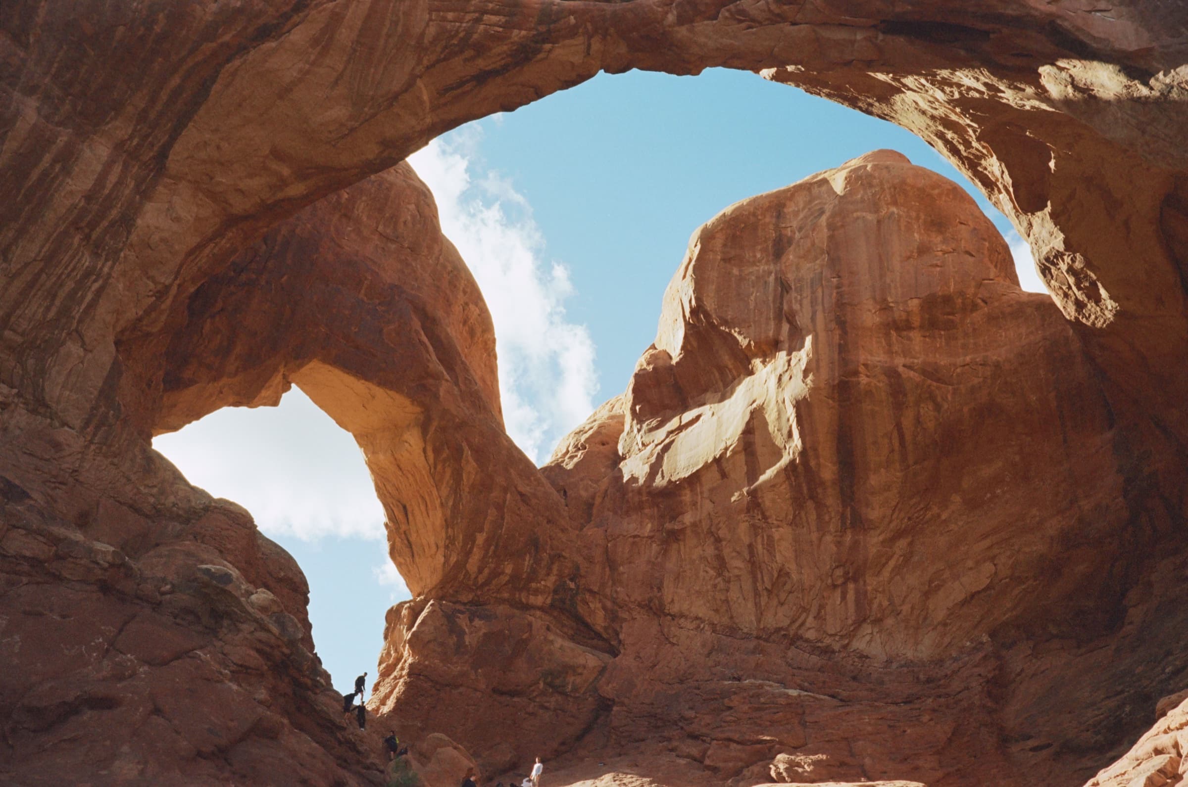 Looking up through the massive sandstone openings of Double Arch at a bright blue sky, a tiny figure below for scale