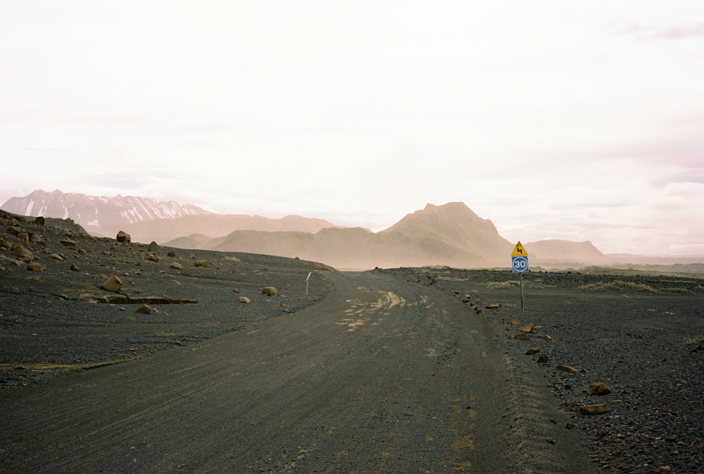 A desolate gravel road cutting through black volcanic highland terrain toward hazy mountains