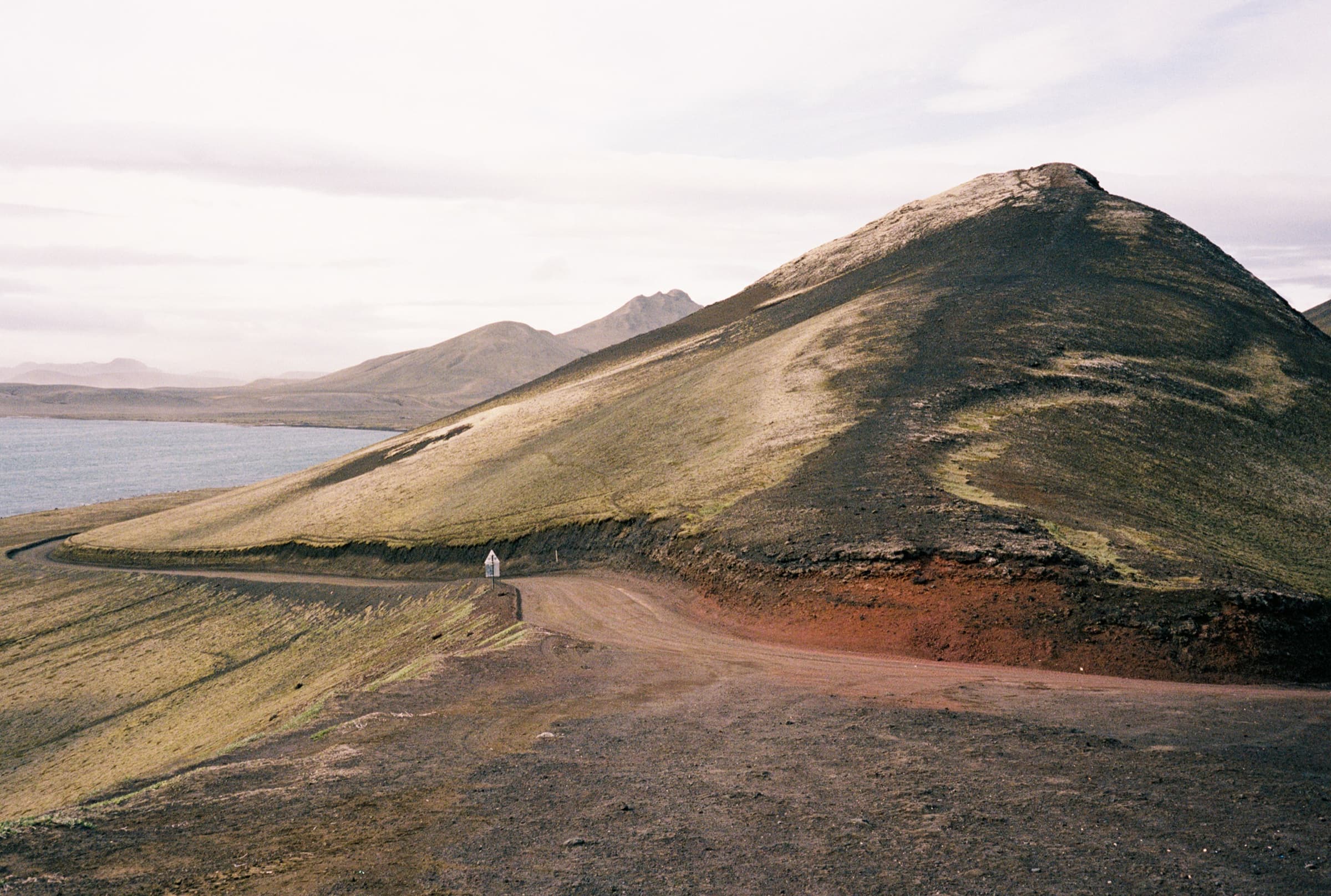 A lone figure on a highland dirt road beside a green volcanic hillside above a glacial lake