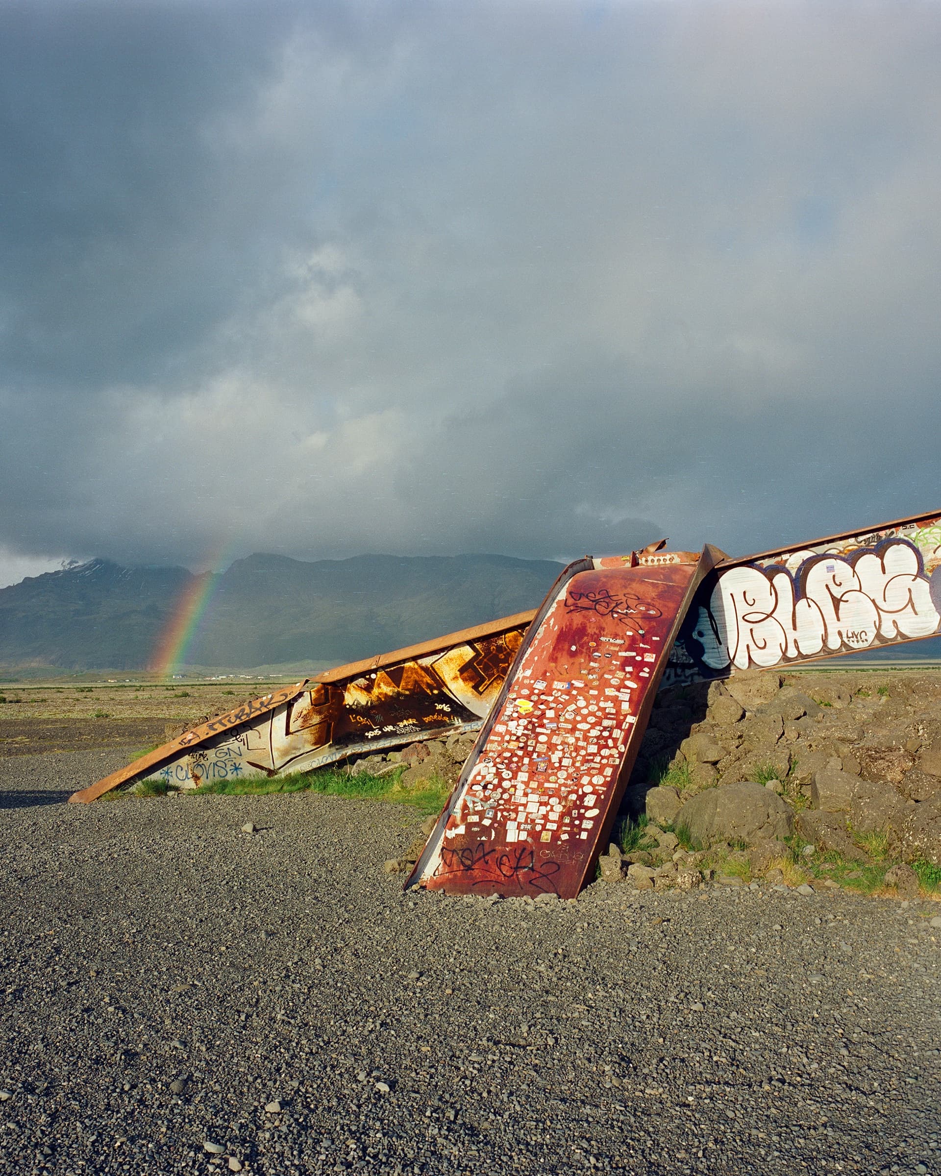 A rusted graffiti-covered bridge girder swept away by a glacial flood, with a rainbow arching in the sky behind it