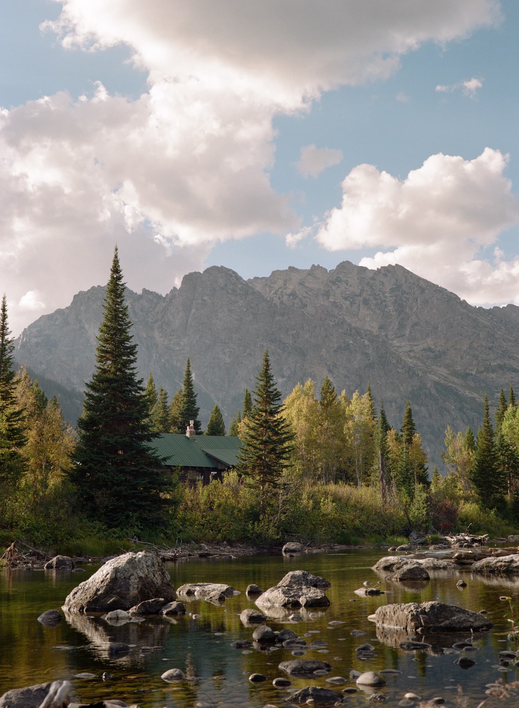A rustic boathouse nestled among pines with the Grand Teton peaks rising above a rocky stream