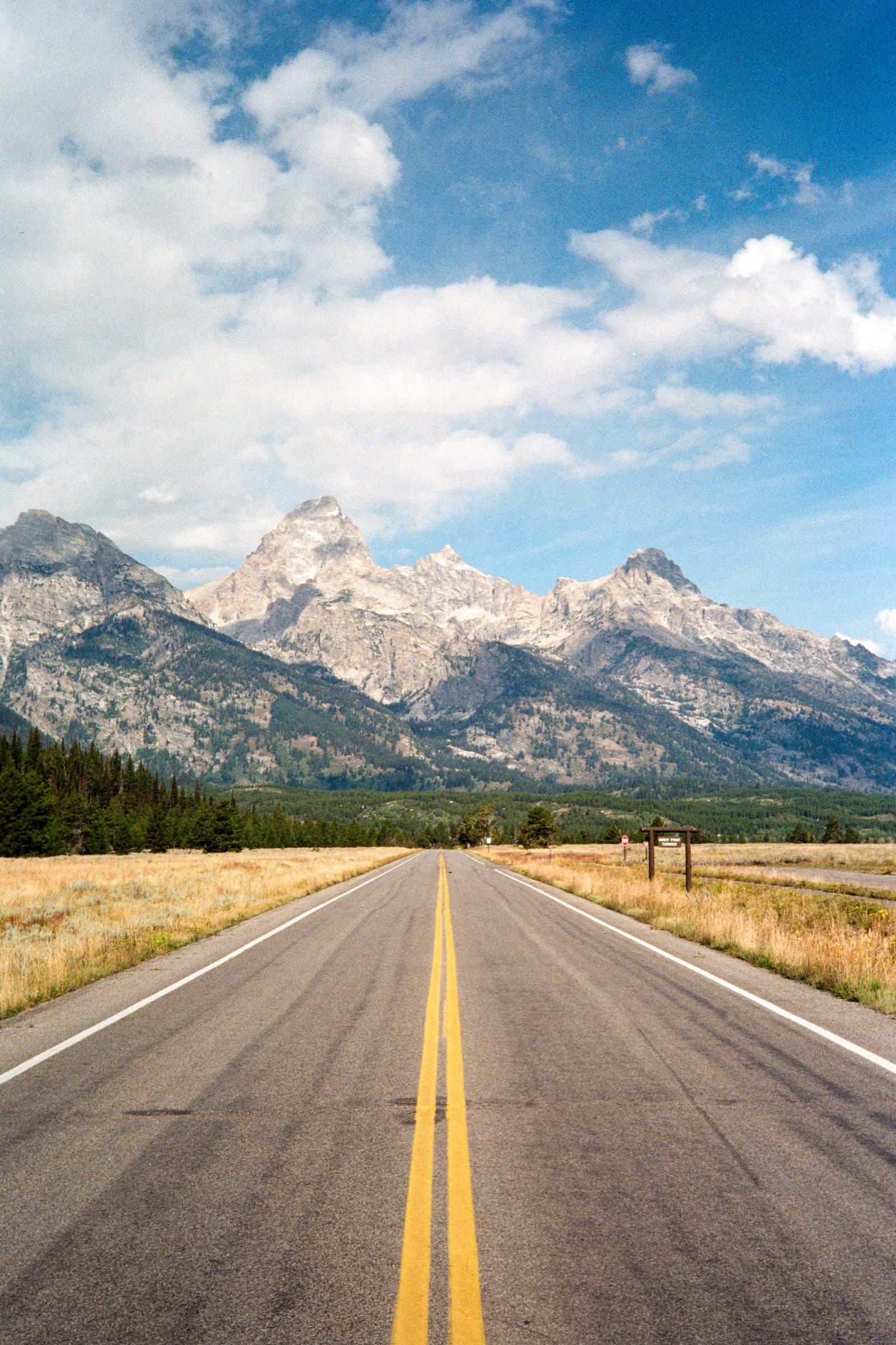 A straight highway leading directly toward the snow-capped Grand Teton peaks through golden prairie